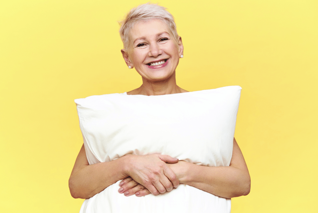 Femme souriante avec un oreiller illustrant les bienfaits du bruit blanc et du bruit rose pour améliorer le sommeil après 60 ans